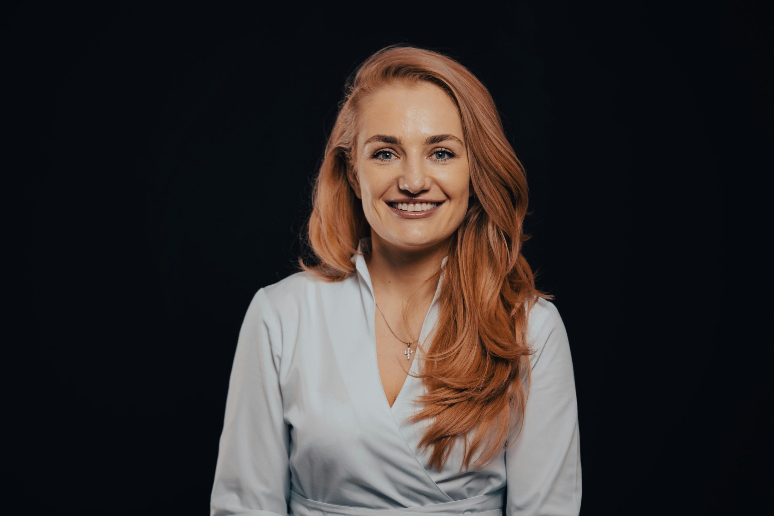 Studio portrait of a smiling woman with red hair in an elegant white dress against a dark background.