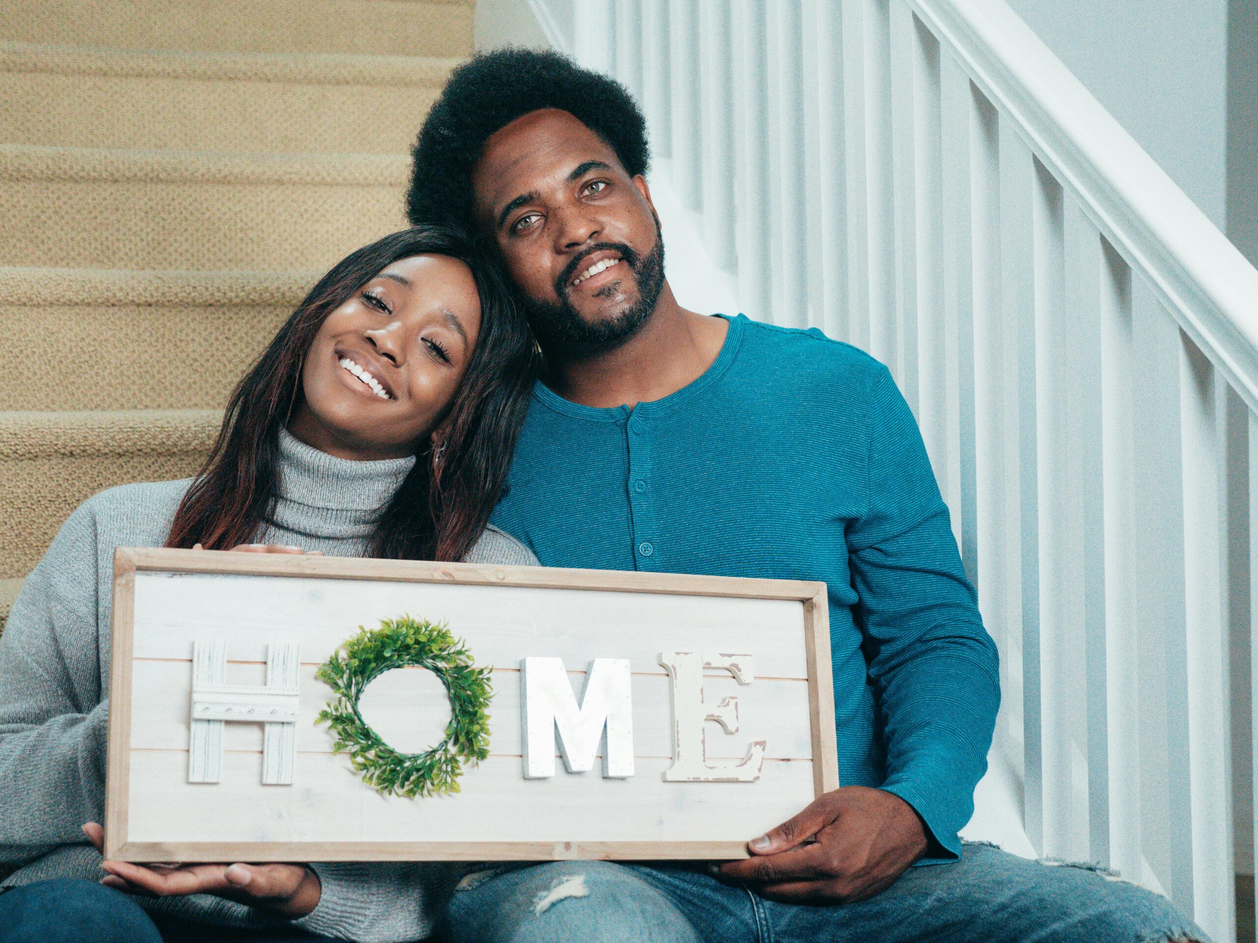 Smiling couple sitting on stairs holding a HOME sign, symbolizing new beginnings in their new house.