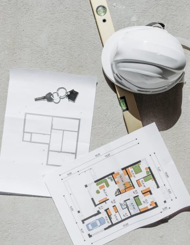 Flat lay of construction plans, keys, hard hat, and level tool on concrete.